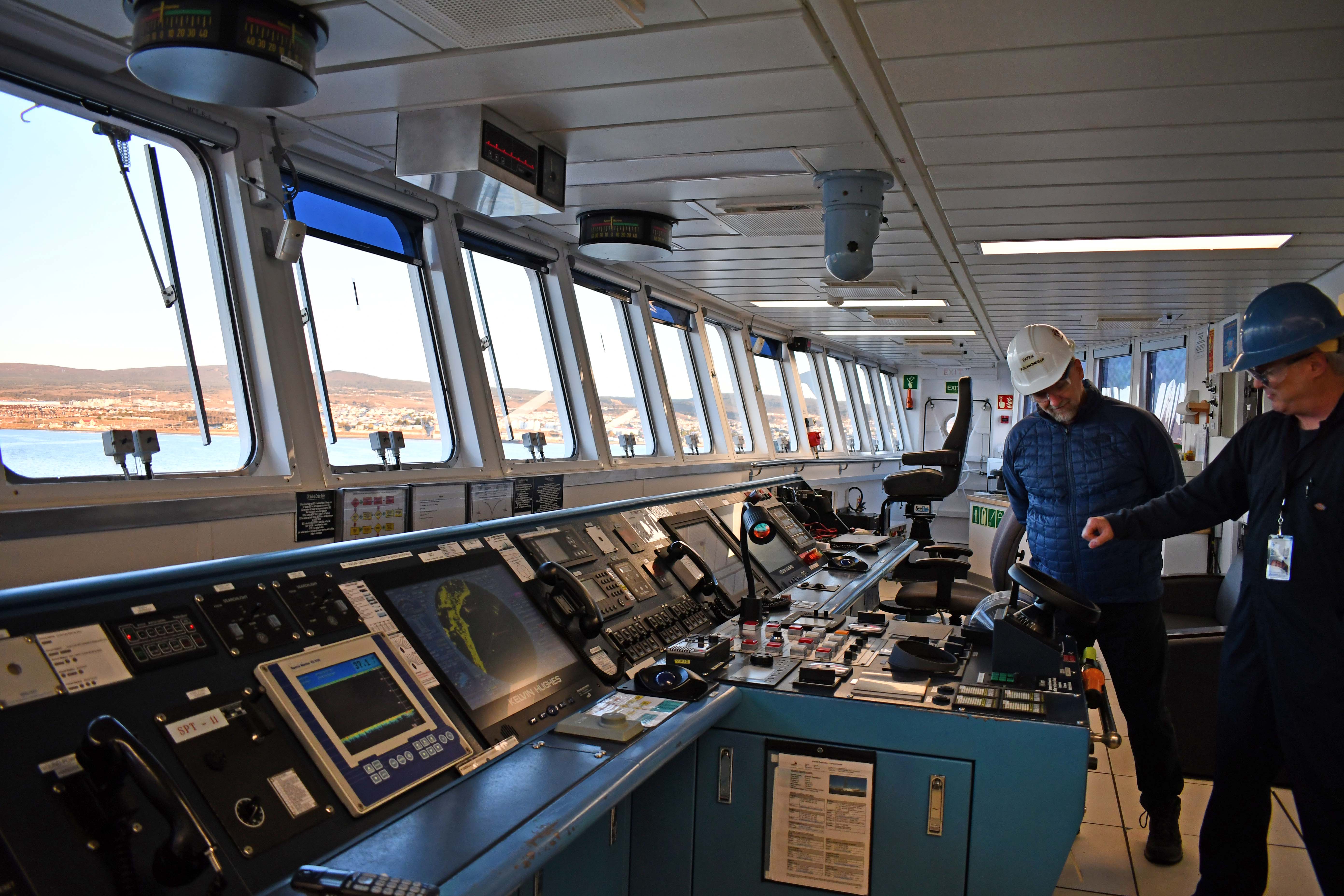 Two men in hardhats look at the instruments on the ship's bridge