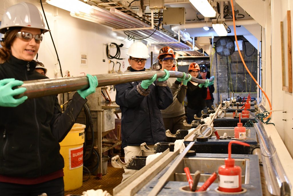 Technicians in hardhats and safety glasses carry a 10 meter sediment core onto the catwalk