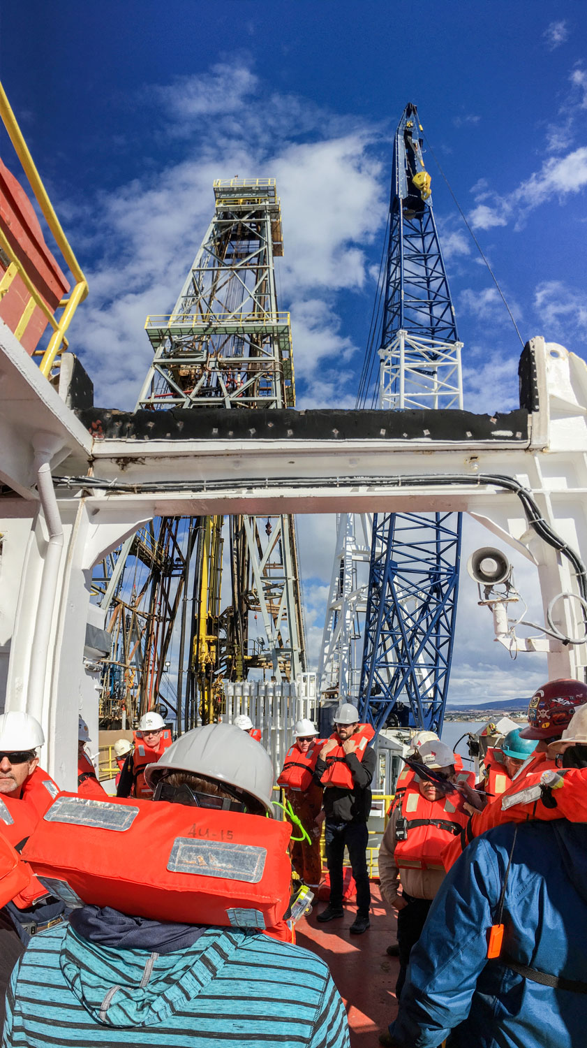 People wearing lifejackets, hardhats, and safety glasses. The derrick in the background.