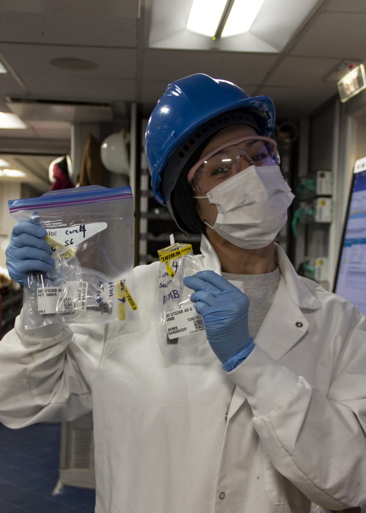 Dr. Linda Armbrecht in a hardhat, safety glasses, face mask, gloves, and lab coat holds up ziploc bags of samples.