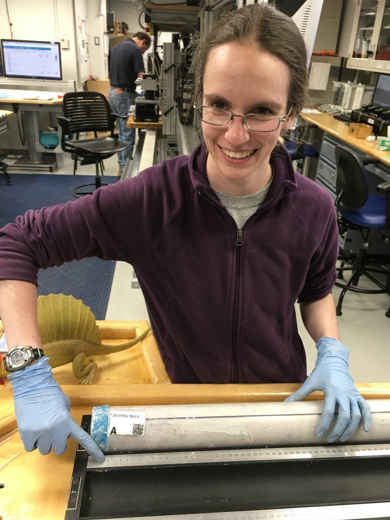 Elizabeth, a short, thin white woman in a purple jacket and glasses, standing next to a section of deep-sea mud in a plastic tube on a table with a ruler next to it. She is pointing at the label and the ruler next to the core and grinning. The core label says Core 15H, Section 5, and her finger is at 3cm, or 15-5-3, just like the site number.