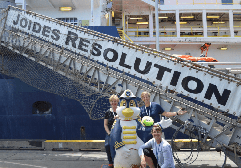 Three women posing next to a penguin mascot. The ship's gangway in the back reads JOIDES RESOLUTION.