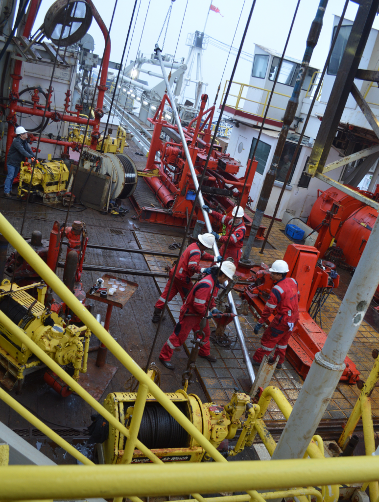 Four people in red coveralls hold a thin, very long metal tube, diagonally pointing toward the ground. There is heavy drilling machinery all around them.