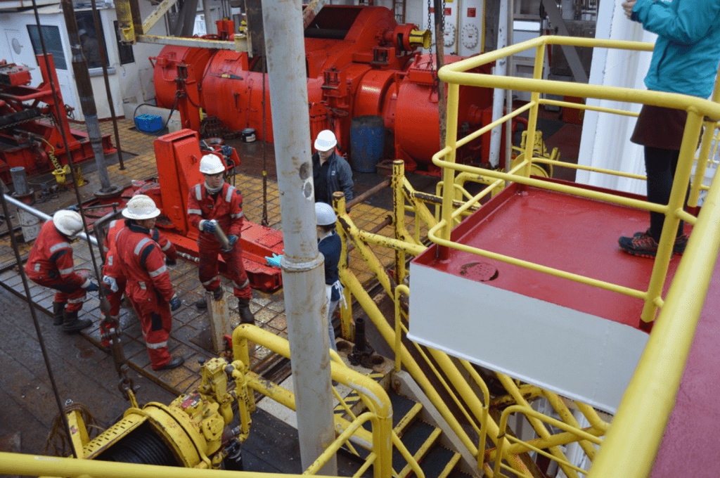 A driller in red coveralls hands a small metal tube, the core catcher, to a lab tech. There are several other people standing with them on the rig floor.