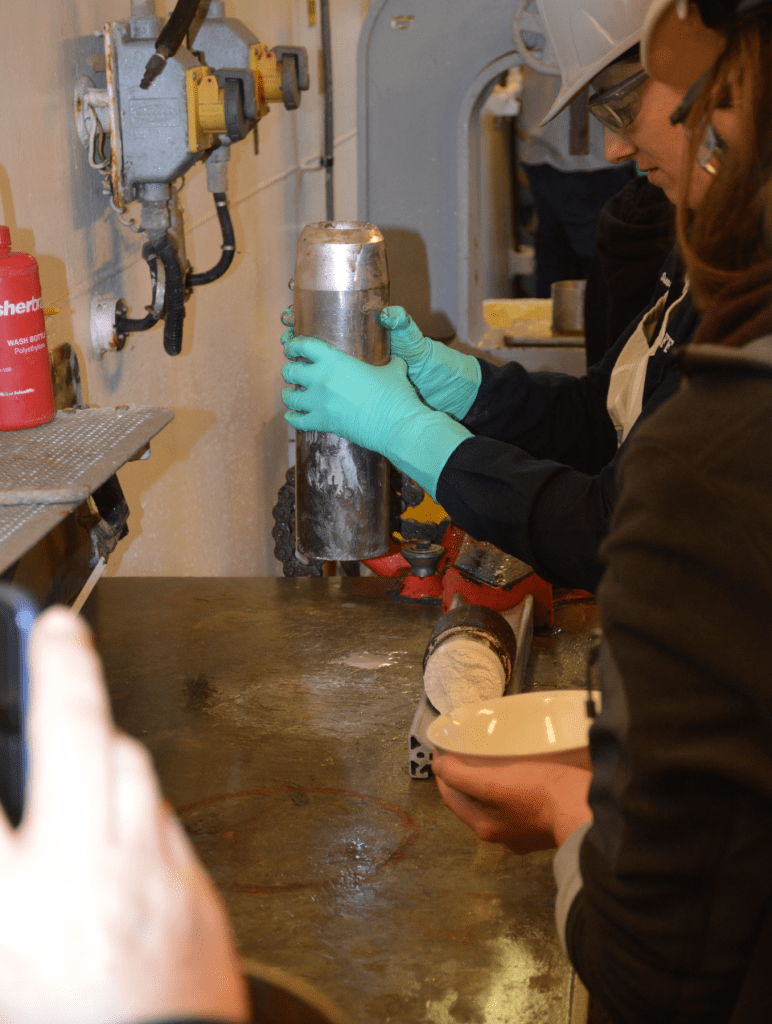 A tech holds the core catcher, a short metal tube, in the air. A scientist waits slightly out of frame with a white ceramic bowl.