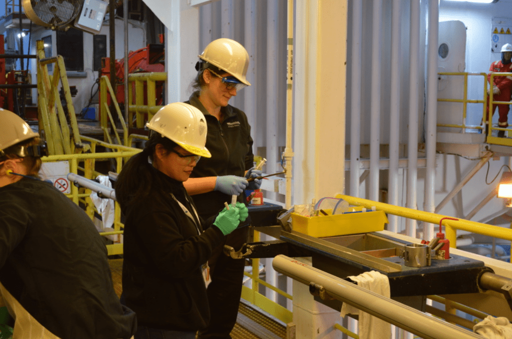 Two scientists stand in front of a core on the catwalk. They are both working with small instruments in their hands.