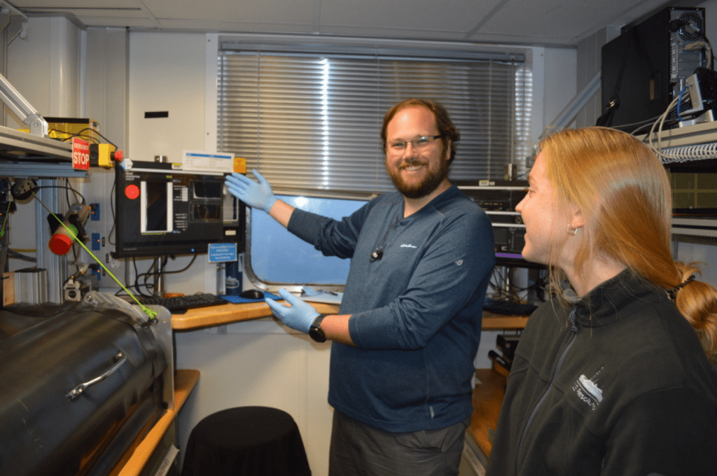 Two scientists standing in front of a machine. One of them is gesturing to the machine while smiling happily, and the other is looking off frame.