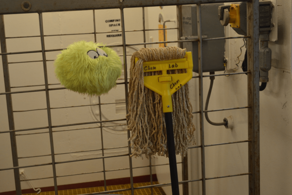 A fuzzy green plushie, Little Cthulhu, sitting on a rack next to a mop head. The mop head looks almost like a person's long hair hanging down. LC is positioned so it looks like it's looking at the mop head.