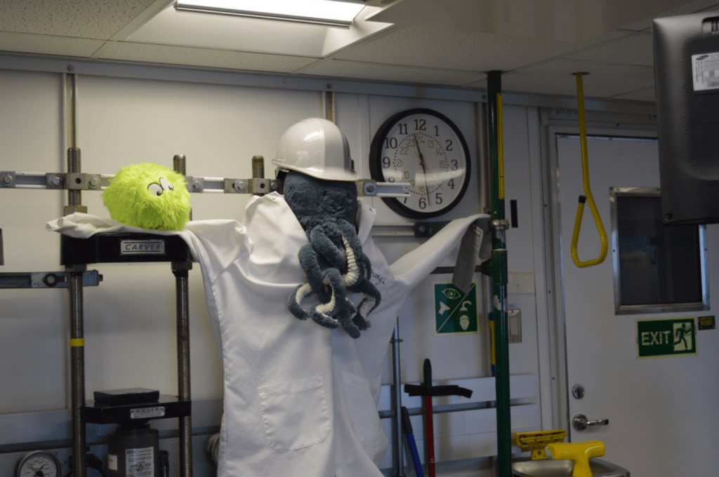 A lab coat hanging from two grey squeeze cake machines. The lab coat has the name "Chang" stitched above the breast pocket. Sitting above the neck hole, as if it was wearing the lab coat, is a blue octopus plushie wearing a hard hat. A fuzzy green plushie, Little Cthulhu, sits on one of the lab coat's arms.