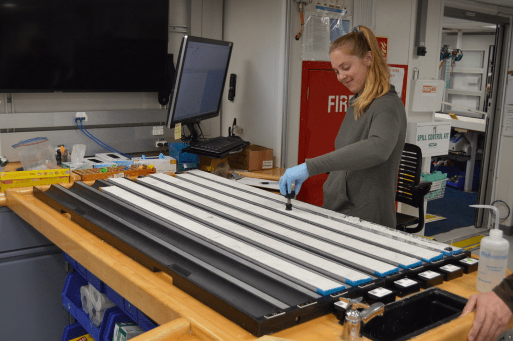 A scientist standing in front of a core workstation. She is pressing a small black instrument into one of the split cores with a slight smile on her face.