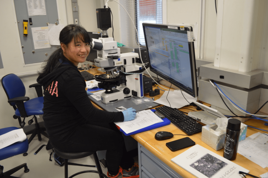 A scientist sitting at a microscope. Her hand is holding a pen over a piece of paper, and she is turned to the camera and smiling. There is a computer monitor with several spreadsheets open above her work station.