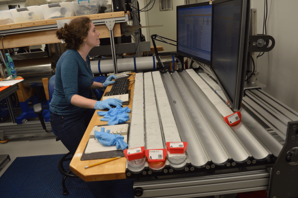 A scientist sitting at a core table. There are several sections of split core sitting on the table in U-shaped channels. She is looking over the table at a computer monitor with a focused expression on her face.