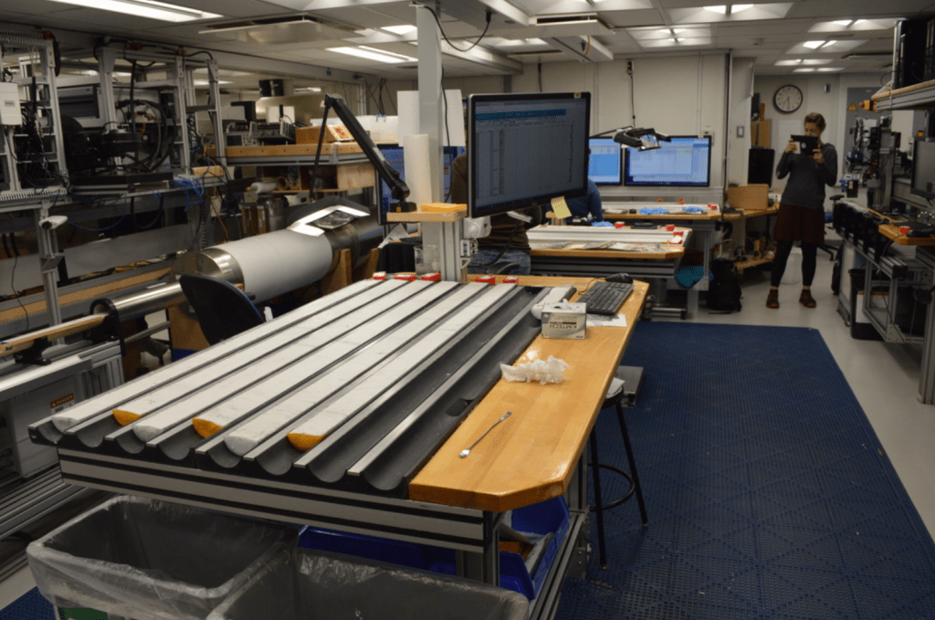 A shot of the core lab. In front of the camera is a table with U-shaped channels mounted into it and a computer mounted nearby. Most of the channels are holding split cores. There are two more of these stations further back in the shot. At the back of the shot is a woman holding an iPad to take a picture. There is a long instrument on a moving track to her left.