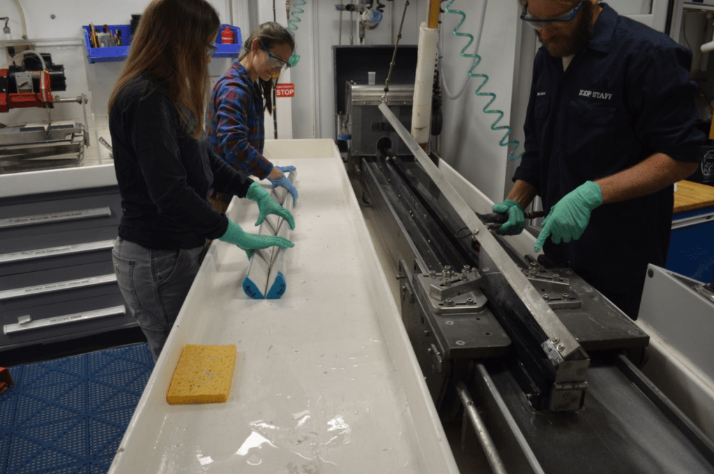 Three lab technicians in the cutting room. One is spraying the cutting table from the last photo with a hose, and the other two are holding a freshly split core on the table. All three people have neutral expressions on their faces.