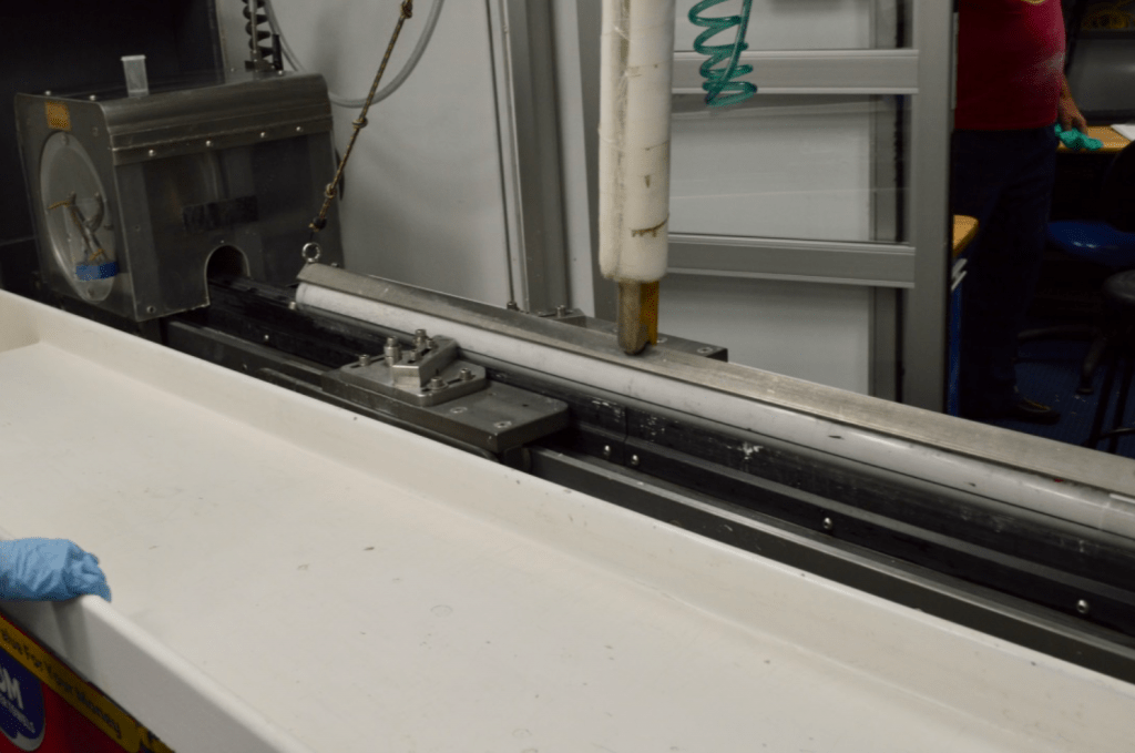 A photo of a machine atop a cutting table. There is a grey box with a hole in the bottom center, and aligned with that hole is a white sediment core in its core liner. A long bar holds the core to the cutting table and a tech's blue gloved hand can be seen at the edge of the picture, lightly gripping the table.