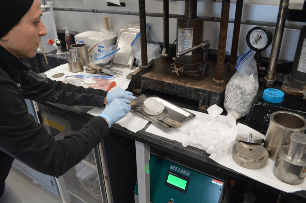 A scientist is touching a squeeze cake with a metal spatula while it sits on a plate. The squeeze cake is a thick disc of sediment, about palm sized. There are several large grey instruments in the background - hydraulic squeezers.