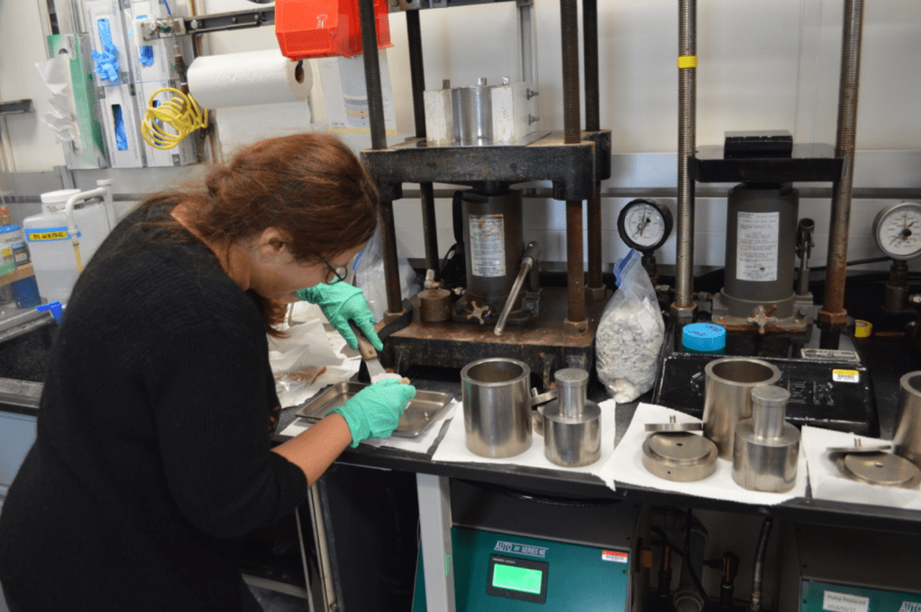 A scientist manipulates a squeeze cake using two metal spatulas. Her back is to the camera and she is working intently. She is surrounded by hydraulic squeezers and large metal implements.