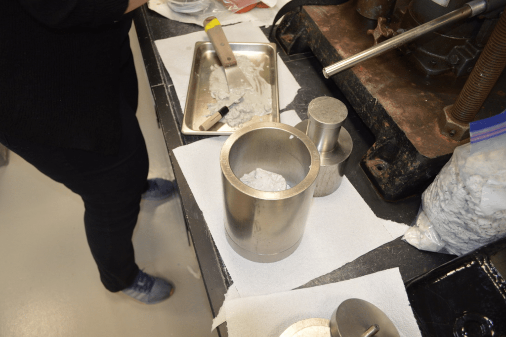 A deep metal cylinder on a lab bench. A white sediment sample is barely visible beneath the rim of the cylinder. There is a muddy plate and metal spatula next to the cylinder - where the sample came from.