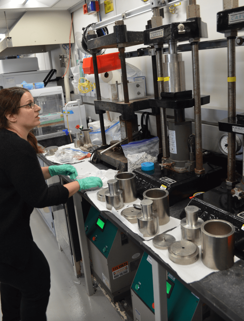 A scientist, Eleni, is looking up intently at the hydraulic squeezer. There is a metal cylinder and a plunger in the jaws of the squeezer. 