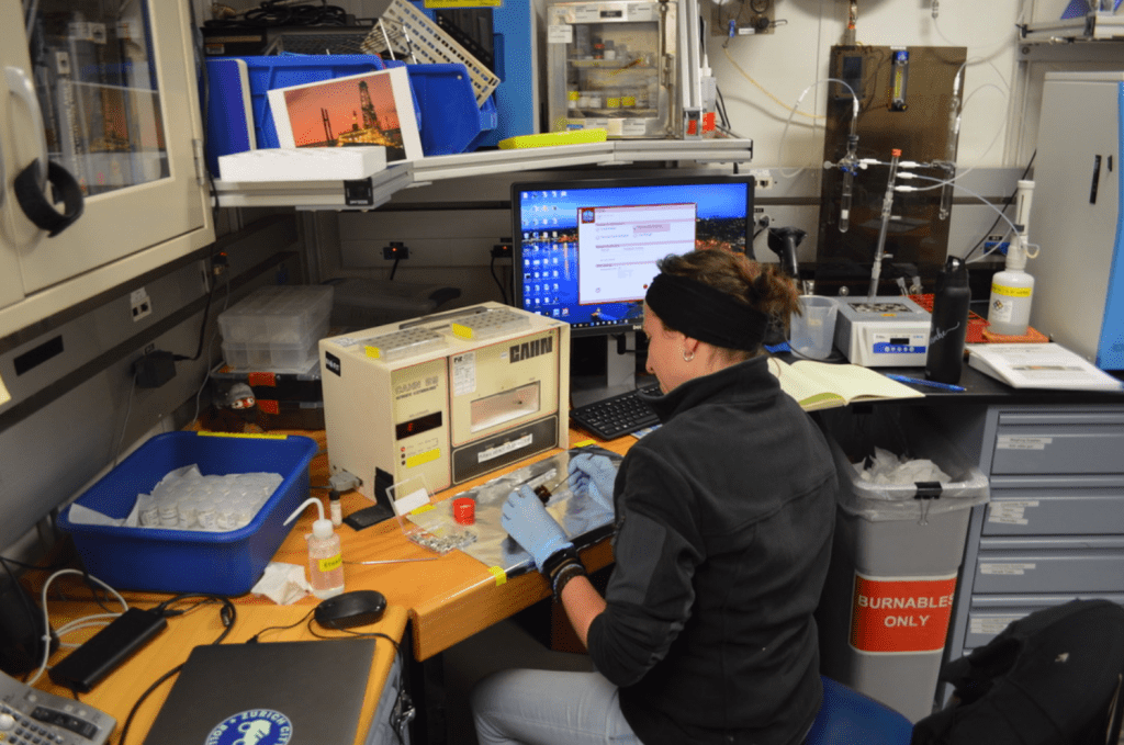 A scientist, Blanca, sits at a lab bench full of machinery. She is scooping a sample from a glass vial.  Her back is turned to the camera.