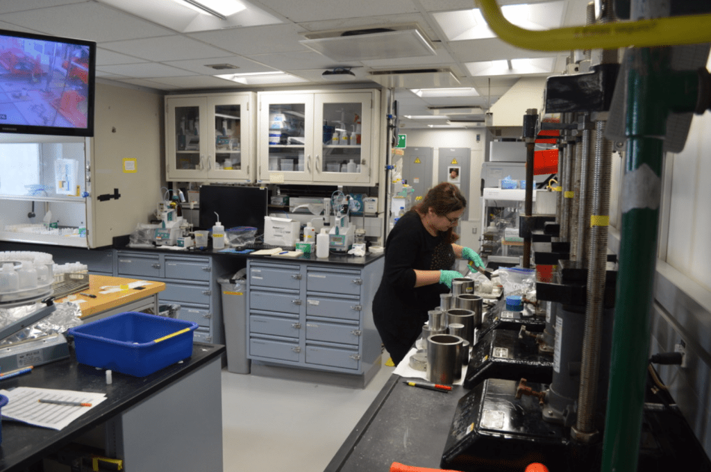 A wide shot of the lab. Eleni is standing at a lab bench lined with hydraulic squeezers. Behind her is a lab bench full of machines with glass tubes sticking out of the tops. There is a television mounted to the wall, and it is showing a grainy grey and red image of the rig floor.