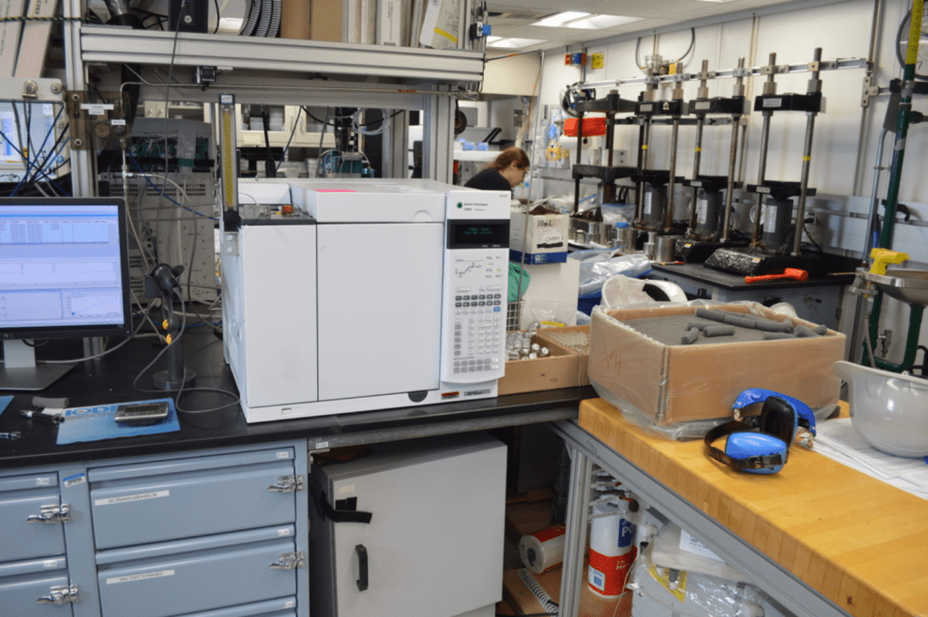 A large (mini-fridge sized) white instrument shaped like a box, sitting on a lab bench. There is a computer next to it and several boxes of glass vials and other chemistry equipment scattered around the lab.