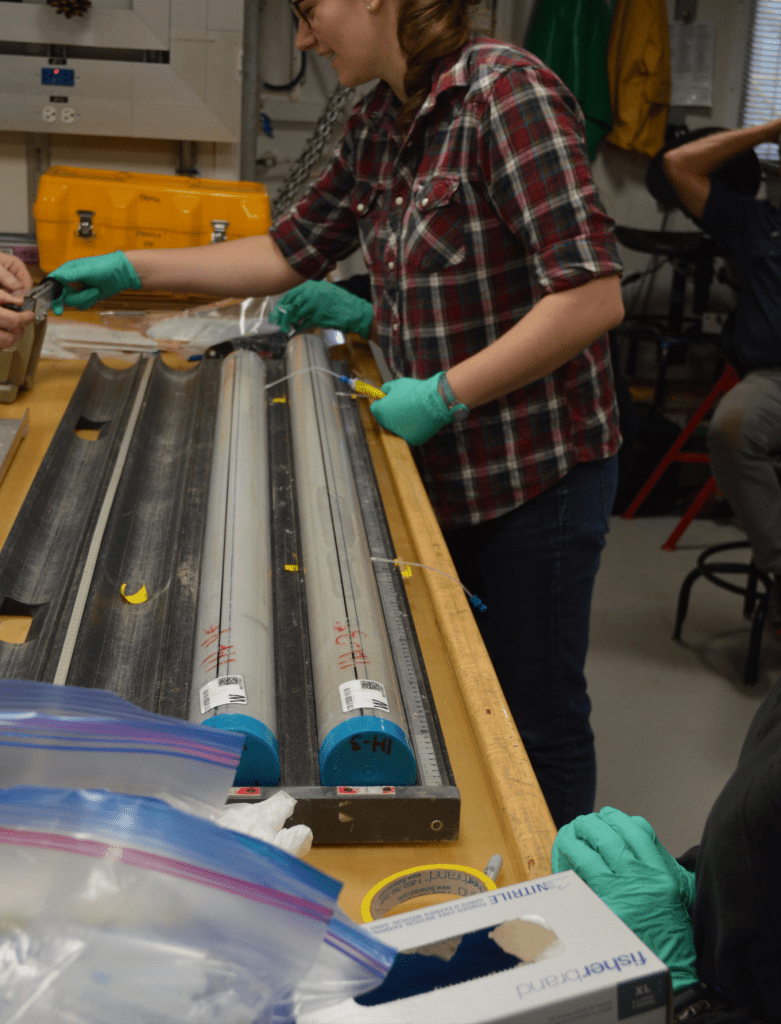 A scientist, Ann, standing at a wooden lab bench. There are two cores in front of her on the bench. Each has a tube in the plastic core liner with a syringe attached to it. Ann is holding one of the syringes and talking to someone off-frame. 