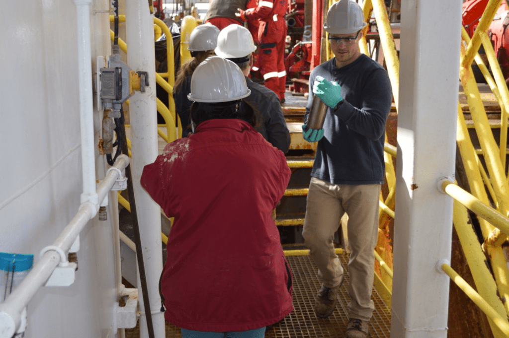 A picture of the catwalk with a few people milling around. A technician, Mark, is carrying a small metal tube in his gloved hands. He is looking down off-frame with a focused expression. In the foreground is another technician, Kenzie, facing away from the camera and wearing a red jacket. The jacket has something white smeared over the left shoulder.