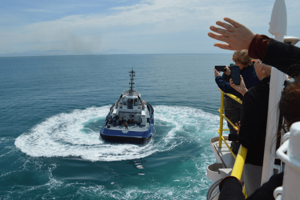 A small tug boat, the Hinewai, within a spiral of its own wake. The ocean is calm and bright blue. The railing of the JR is to the right of the photo, and several people are waving at the boat over the railing and taking pictures.