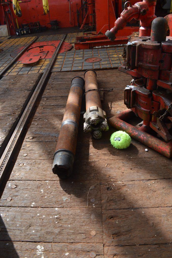 Two weathered-looking pieces of drill pipe, one with a drill bit at the end, sitting on the rig floor. A fuzzy green plushie (Little Cthulhu) sits on the floor next to the drill pipe.