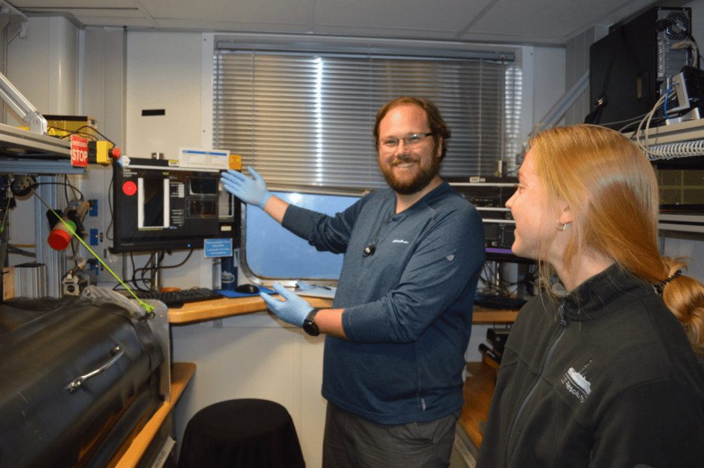 Two scientists standing in a corridor lined with lab equipment. One of the scientists, Alex Reis, is grinning at the camera and gesturing towards a computer screen with data readouts on it. The other scientist, Gabby Kitch, is looking at Alex and smiling.