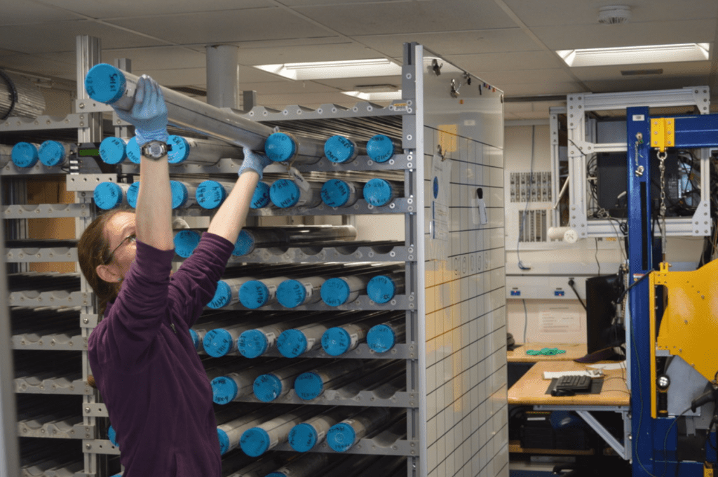 A scientist, Elizabeth Sibert, with her arms above her head, pulling a core out of a rack. She is looking up at the core, which is grey in color.