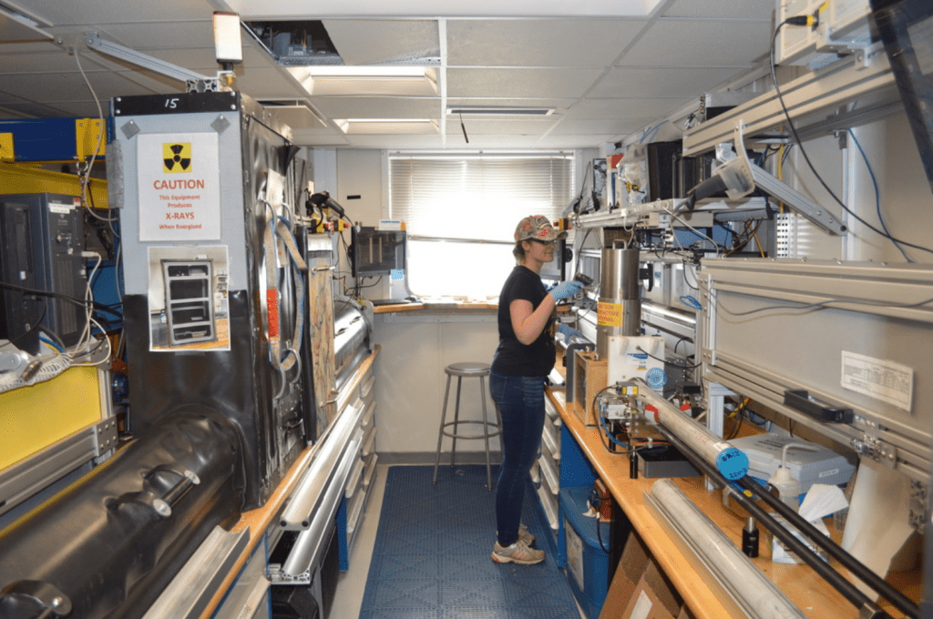 A scientist, Heather Jones, standing in a corridor lined with lab equipment. She is using a handheld barcode reader to scan something we can't see. There is a neutral expression on her face.