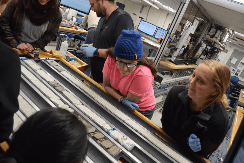 A few scientists standing around a table with core halves set on it. A scientist, Gabby Kitch, is looking at someone off-frame with a slight smile. Another scientist, Laura Haynes, is looking down at the core. Alex Reis is slightly in the background, holding his glasses in his hands and looking down. There are a few other scientists partially in frame and looking at the core.