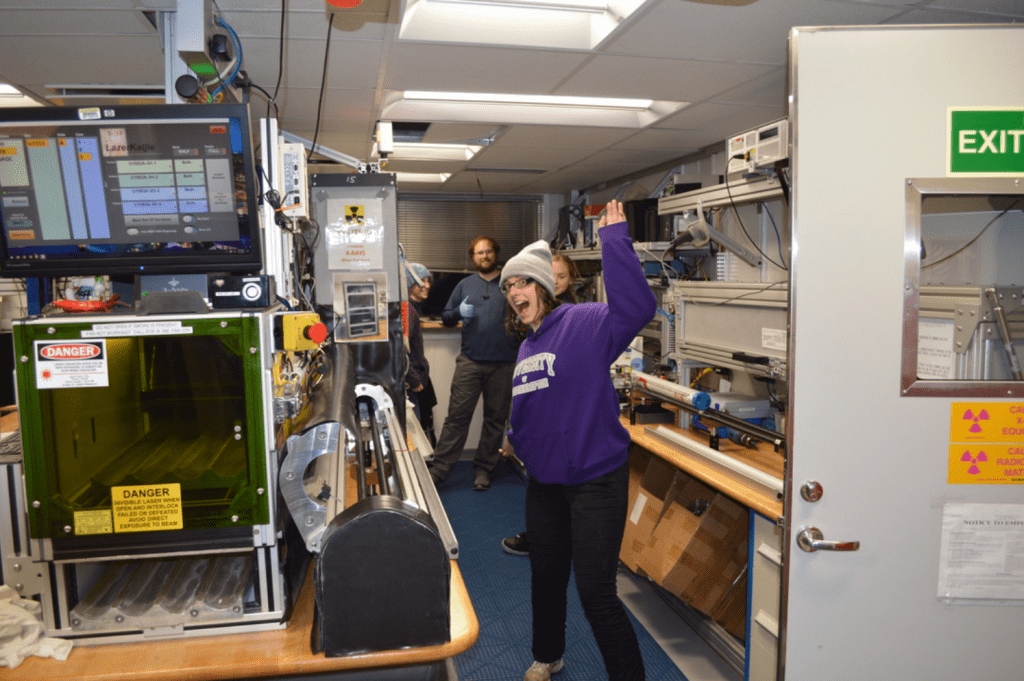 The physical properties corridor of the lab, lined with lab equipment. Alex, Gabby, and Elizabeth are in the background and smiling at the camera. Alex is giving the camera a thumbs up. Heather Jones is in the foreground, waving to the camera with a large, open-mouthed smile on her face.