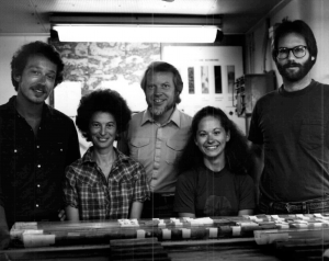 A black and white photo of five scientists standing at a table with core on it. The scientists are all looking at the camera and smiling.