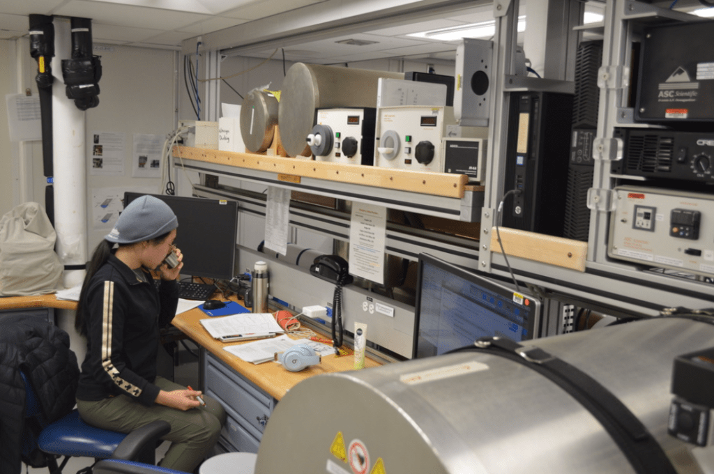 A scientist, Wendy Zhang, sits at a table lined with lab equipment and computer screens. She is turned away from the camera and is sipping coffee out of a small cup. The lab bench is partially obscured by a large, silver cylinder - a magnetometer.