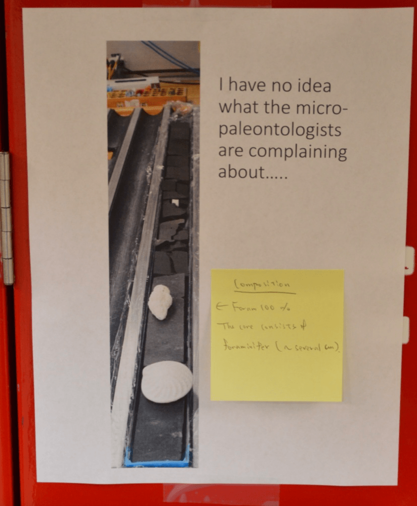 A printed-out picture taped to a red wall. The picture is of a dark grey core with a couple bright white 3D-printed forms set on top. The caption of the picture is "I have no idea what the micro-paleontologists are complaining about..." There is a sticky note attached to the picture that says "Composition: Foram 100%. The core consists of foraminifer (~several centimeters)."