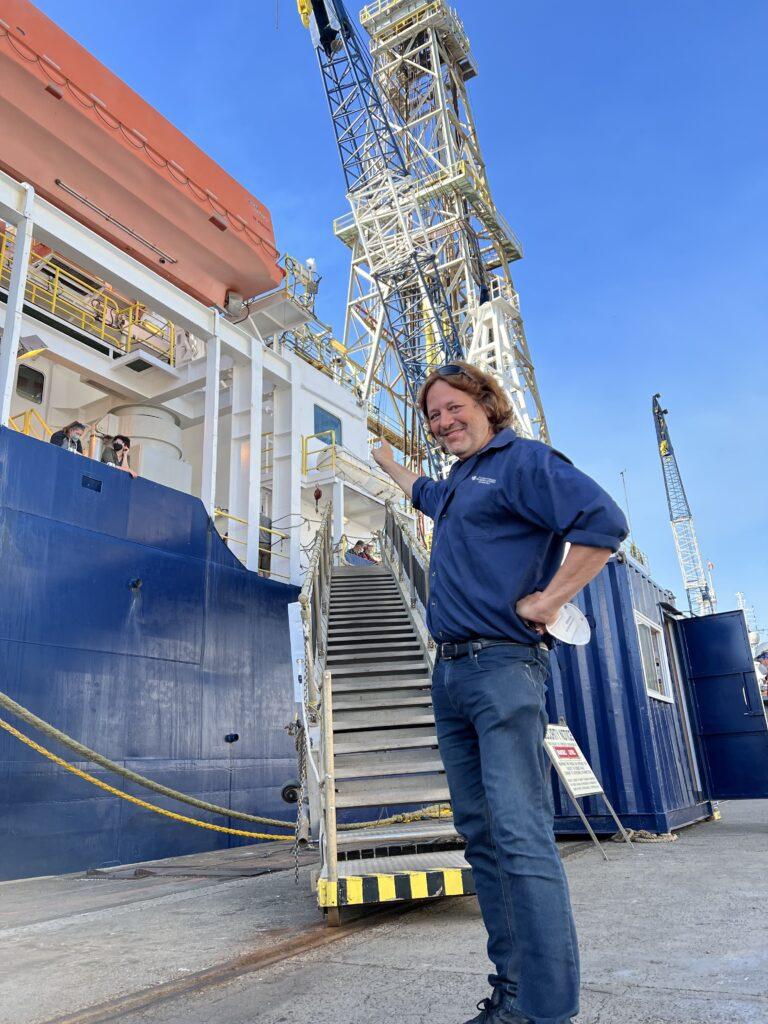 A man standing in front of stairs that lead up to a large ship. He is pointing at the ship.