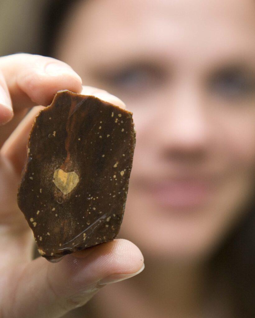 A person blurred in the background holding up a small rock in between their fingers that has a heart shaped spot inside it.