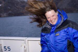 Carol is smiling with her dark blond hair blowing behind her in the wind. She is wearing a blue and black wind breaker. In the background, the sea and steep land are visible.