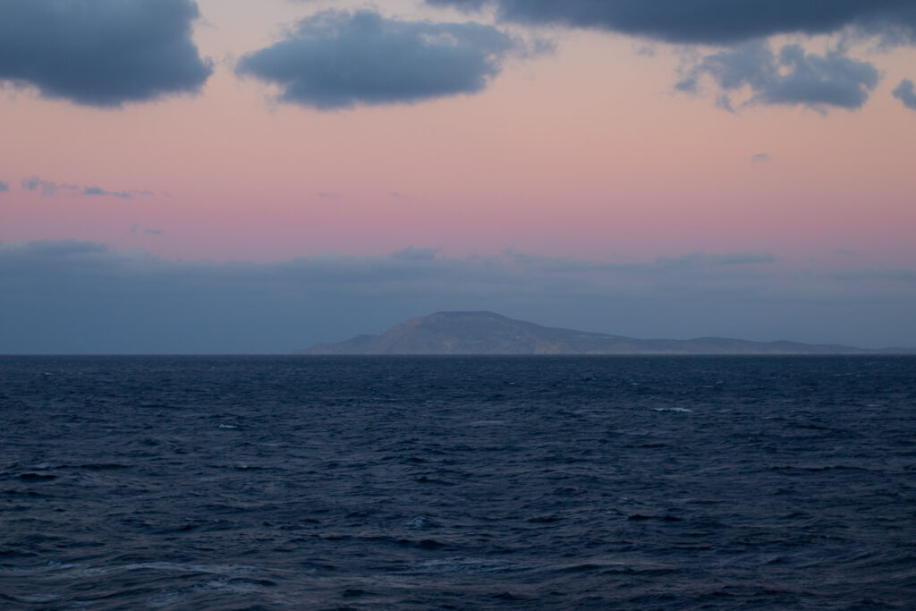 A small island is seen across the water at sunrise. The sky moves from dark blue at the horizon to pink to orange near the clouds.