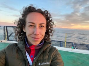 Maya is standing on the green JOIDES Resolution helideck with the setting sun behind her. Her brown curly hair is blown back by the wind. She is smiling. She is wearing an olive green jacket over a purple and red fleece.