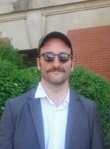 Rob is standing in front of a bush with a brick building in the background.