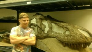 Arthur stands with his arms crossed in front of a dinosaur skull fossil.