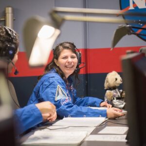 Alejandra is sitting at a desk wearing a headset and a blue jumpsuit.