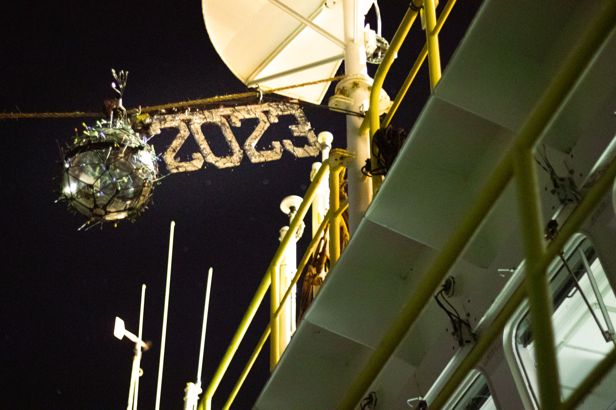A christmas-light covered ball hangs next to decorative 2023 numbers. In the background we see a ship's deck.
