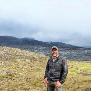 David stands on a grassy field with a black rocky landscape in the background.