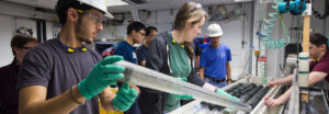 In the foreground, a person wearing rubber gloves and a hard hat holds up a core liner, while several people in the background look on.