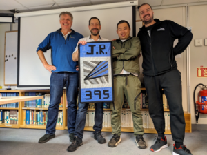 An image of four people standing together and smiling while they hold up a banner that reads J.R. 395